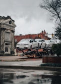 A car transporter is carrying several white luxury cars on an urban street. The background includes a historic building with classical architecture and a traffic light. It's an overcast day with a muted atmosphere.
