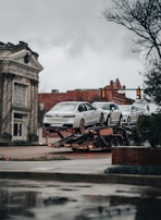 A car transporter is carrying several white luxury cars on an urban street. The background includes a historic building with classical architecture and a traffic light. It's an overcast day with a muted atmosphere.