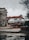 A car transporter is carrying several white luxury cars on an urban street. The background includes a historic building with classical architecture and a traffic light. It's an overcast day with a muted atmosphere.