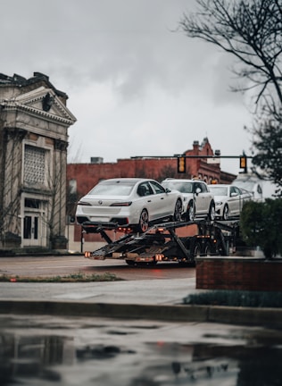 A car transporter is carrying several white luxury cars on an urban street. The background includes a historic building with classical architecture and a traffic light. It's an overcast day with a muted atmosphere.