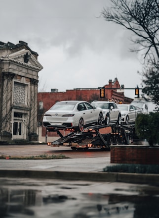 A car transporter is carrying several white luxury cars on an urban street. The background includes a historic building with classical architecture and a traffic light. It's an overcast day with a muted atmosphere.