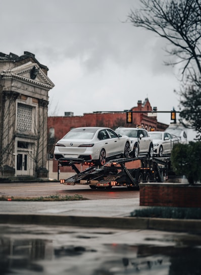 A car transporter is carrying several white luxury cars on an urban street. The background includes a historic building with classical architecture and a traffic light. It's an overcast day with a muted atmosphere.
