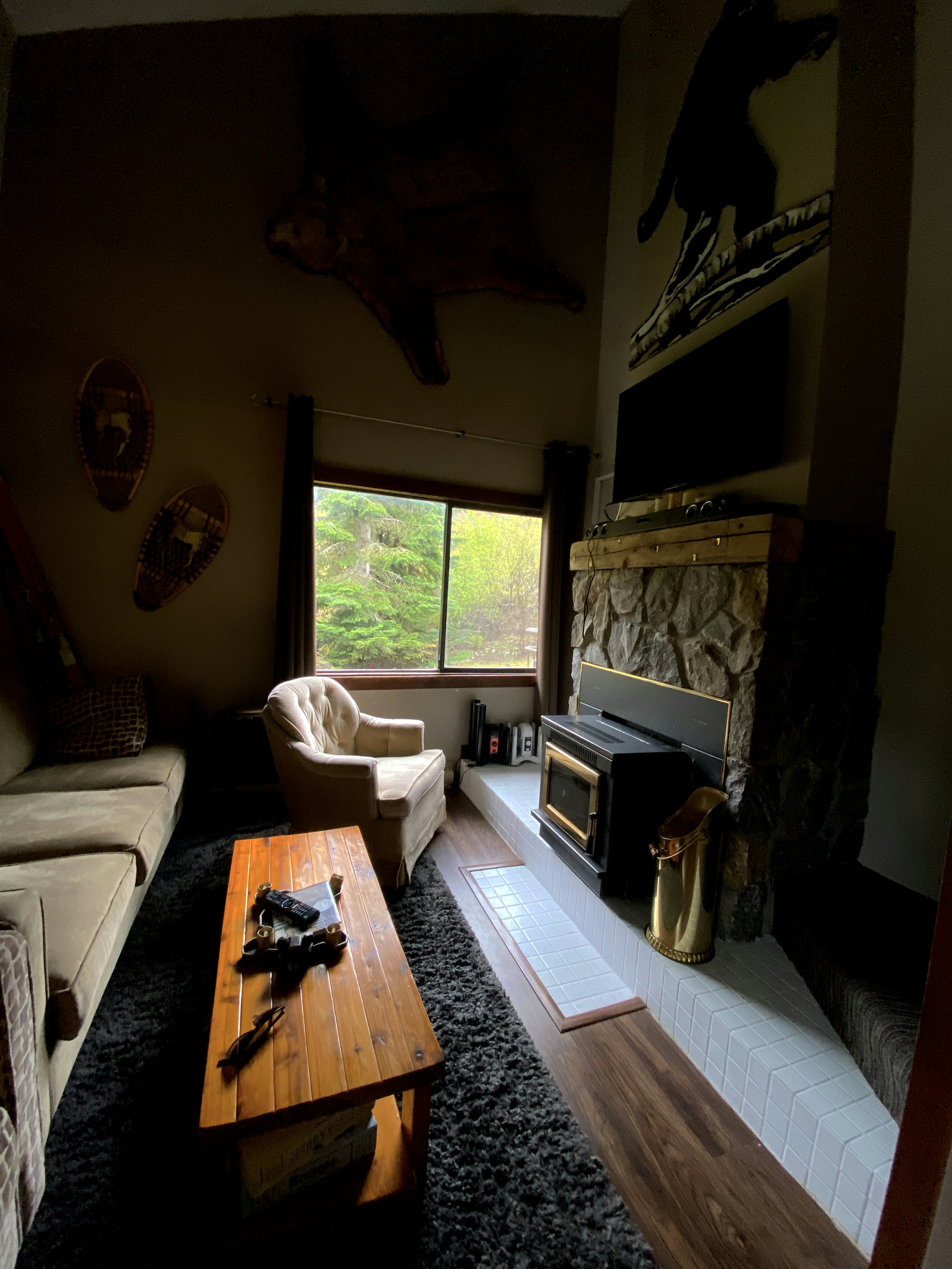 A cozy living room with rustic decor features a plush beige armchair and sofa situated around a wooden coffee table. A fireplace with a stone finish and a metal wood-burning stove adds warmth. Large window allows natural light to enter, overlooking greenery outside. Décor includes mounted snowshoes and a large, mounted animal skin above the window.
