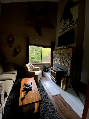 A cozy living room with rustic decor features a plush beige armchair and sofa situated around a wooden coffee table. A fireplace with a stone finish and a metal wood-burning stove adds warmth. Large window allows natural light to enter, overlooking greenery outside. Décor includes mounted snowshoes and a large, mounted animal skin above the window.