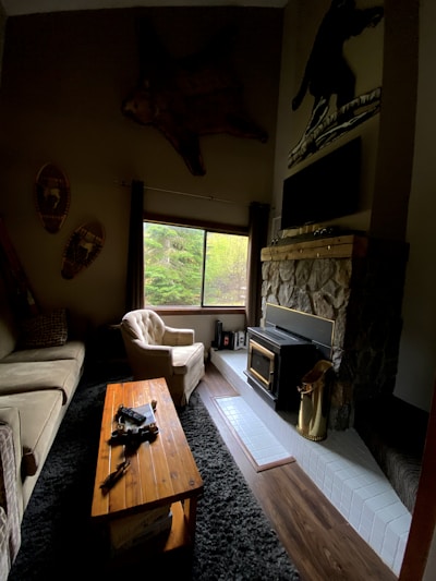 A cozy living room with rustic decor features a plush beige armchair and sofa situated around a wooden coffee table. A fireplace with a stone finish and a metal wood-burning stove adds warmth. Large window allows natural light to enter, overlooking greenery outside. Décor includes mounted snowshoes and a large, mounted animal skin above the window.