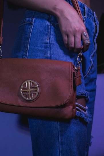 A person wearing denim jeans with a distressed design holds a brown leather bag. The bag features a circular, decorative emblem. The lighting is bluish, adding a moody atmosphere.