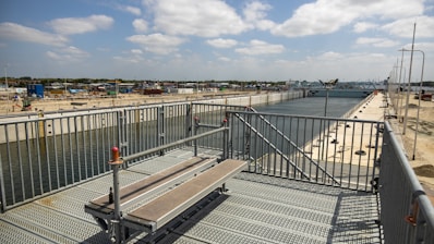 A large industrial canal or waterway stretches into the distance, flanked by metal fencing and construction infrastructure. The sky is partly cloudy, and there are fenced-off areas and construction equipment visible. The scene suggests an ongoing building or engineering project.
