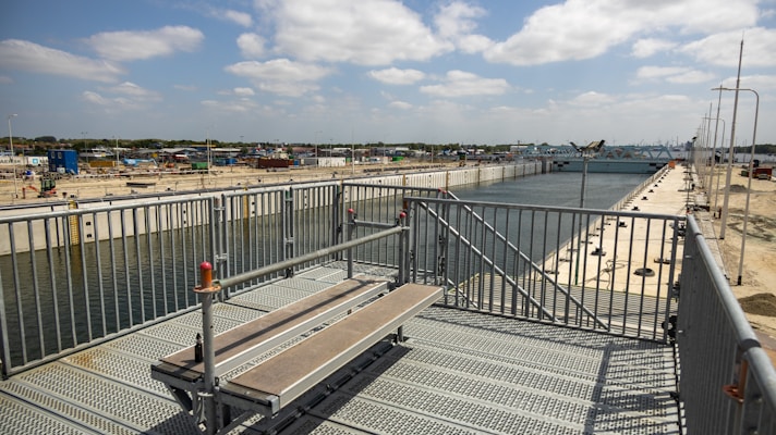 A large industrial canal or waterway stretches into the distance, flanked by metal fencing and construction infrastructure. The sky is partly cloudy, and there are fenced-off areas and construction equipment visible. The scene suggests an ongoing building or engineering project.