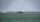 A large cargo ship is sailing on a calm body of water. In the background, there are wind turbines on a stretch of land, indicating a focus on renewable energy. The sky is partly cloudy, and there is a forested area visible along the coast.