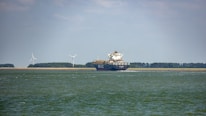 A large cargo ship is sailing on a calm body of water. In the background, there are wind turbines on a stretch of land, indicating a focus on renewable energy. The sky is partly cloudy, and there is a forested area visible along the coast.