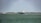 A large cargo ship is sailing on a calm body of water. In the background, there are wind turbines on a stretch of land, indicating a focus on renewable energy. The sky is partly cloudy, and there is a forested area visible along the coast.