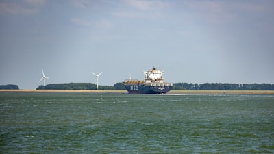 A large cargo ship is sailing on a calm body of water. In the background, there are wind turbines on a stretch of land, indicating a focus on renewable energy. The sky is partly cloudy, and there is a forested area visible along the coast.