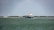 A large cargo ship is sailing on a calm body of water. In the background, there are wind turbines on a stretch of land, indicating a focus on renewable energy. The sky is partly cloudy, and there is a forested area visible along the coast.