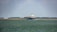 A large cargo ship is sailing on a calm body of water. In the background, there are wind turbines on a stretch of land, indicating a focus on renewable energy. The sky is partly cloudy, and there is a forested area visible along the coast.
