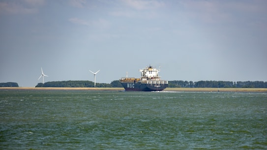 A large cargo ship is sailing on a calm body of water. In the background, there are wind turbines on a stretch of land, indicating a focus on renewable energy. The sky is partly cloudy, and there is a forested area visible along the coast.