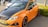 A parked orange car is prominently featured, with a sporty design and black accents including the roof and wheels. The license plate reads S-277-PB. The car is parked on a paved area with a small green hedge nearby. In the background, parts of other vehicles and blurred figures are visible.