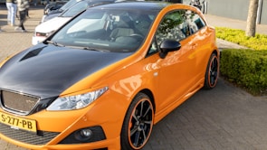 A parked orange car is prominently featured, with a sporty design and black accents including the roof and wheels. The license plate reads S-277-PB. The car is parked on a paved area with a small green hedge nearby. In the background, parts of other vehicles and blurred figures are visible.