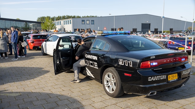 A black police car with 'CANTON 911' markings is parked on a paved area. A person is standing next to the open door of a white car nearby. Several other cars and a group of people are present in the background. The scene takes place in an open area with modern buildings and greenery.