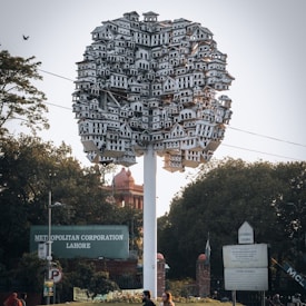 An artistic structure resembling a tree, composed of countless small, white, intricate house models clustered together atop a white pole. Surrounding the structure are green trees and a few buildings. Signs indicate locations in Lahore, Pakistan.