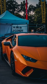 An orange sports car with sleek, angular lines is parked in front of a blue tent. The background includes tall trees and promotional banners with text. The car has a striking, glossy finish and sporty design, featuring aerodynamic features and black rims.