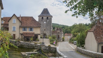 A quaint village scene features a stone building with a clock tower, surrounded by residential houses with red-tiled roofs. A narrow street winds through the village, lined with stone walls and lush greenery. The background includes forested hills under a partly cloudy sky, indicating a peaceful rural setting.