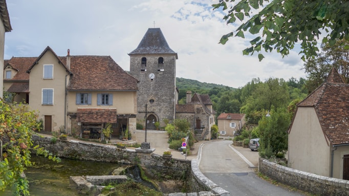A quaint village scene features a stone building with a clock tower, surrounded by residential houses with red-tiled roofs. A narrow street winds through the village, lined with stone walls and lush greenery. The background includes forested hills under a partly cloudy sky, indicating a peaceful rural setting.
