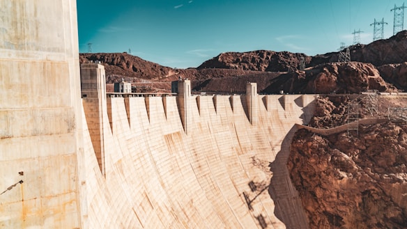 A large concrete dam stretches across a deep canyon with rocky terrain surrounding it. The structure is set against a clear blue sky, and power lines with pylons are visible in the background.