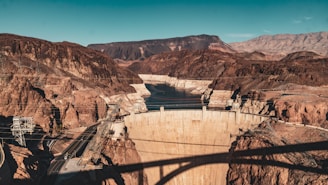 A massive concrete dam set in a rocky canyon with surrounding mountainous terrain. A body of water is visible behind the dam, and there are several bridges and power lines crossing the area. The landscape is arid, with reddish-brown earth tones dominating the scene.