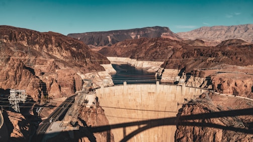A massive concrete dam set in a rocky canyon with surrounding mountainous terrain. A body of water is visible behind the dam, and there are several bridges and power lines crossing the area. The landscape is arid, with reddish-brown earth tones dominating the scene.