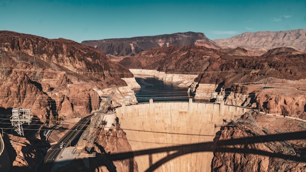 A massive concrete dam set in a rocky canyon with surrounding mountainous terrain. A body of water is visible behind the dam, and there are several bridges and power lines crossing the area. The landscape is arid, with reddish-brown earth tones dominating the scene.