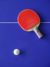 A red table tennis paddle is placed on a blue table tennis table with a white ball nearby. The table has a white line dividing it.