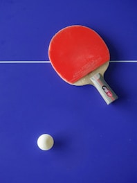A red table tennis paddle is placed on a blue table tennis table with a white ball nearby. The table has a white line dividing it.