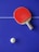 A red table tennis paddle is placed on a blue table tennis table with a white ball nearby. The table has a white line dividing it.