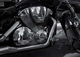A close-up of a motorcycle engine with chrome parts reflecting the surrounding cityscape, including buildings and pedestrians.