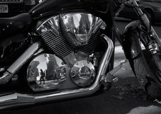 A close-up of a motorcycle engine with chrome parts reflecting the surrounding cityscape, including buildings and pedestrians.