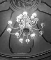 A large, ornate chandelier hangs from an intricately designed ceiling. The chandelier features multiple arms with decorative elements and lights at the end of each arm, casting a warm glow. The ceiling has detailed patterns and curved designs framing the chandelier.