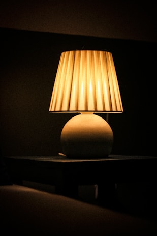 A warm, soft glow emanates from a table lamp with a pleated lampshade. The lamp sits on a dark table against a dimly lit background, creating a cozy and intimate atmosphere.