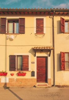 A charming, small yellow house with a rustic wooden door and windows featuring closed wooden shutters. Bright red flower boxes adorn the lower windowsills, adding a touch of color to the exterior. The roof has terracotta tiles, and the warm lighting creates a serene, inviting atmosphere.