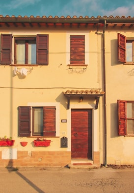 A charming, small yellow house with a rustic wooden door and windows featuring closed wooden shutters. Bright red flower boxes adorn the lower windowsills, adding a touch of color to the exterior. The roof has terracotta tiles, and the warm lighting creates a serene, inviting atmosphere.