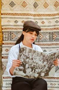 A person with black hair is wearing a vintage-style flat cap and a white shirt, sitting in front of a patterned tapestry, engrossed in reading a newspaper printed in Arabic script. The hanging tapestry features traditional geometric designs in black, brown, and beige.