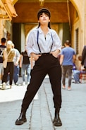 A person stands confidently in a fashionable pose, wearing a white button-up shirt with suspenders, black high-waisted trousers, and black boots. The backdrop is an urban setting, with people walking in the background through an arched walkway. Warm natural lighting highlights the person's expressive stance.