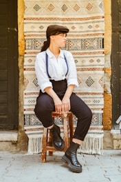 A person in vintage-style clothing, including a white shirt, black suspenders, and black boots, sits on a wooden stool against a decorative tapestry with geometric patterns. The setting appears rustic with textured stone walls.