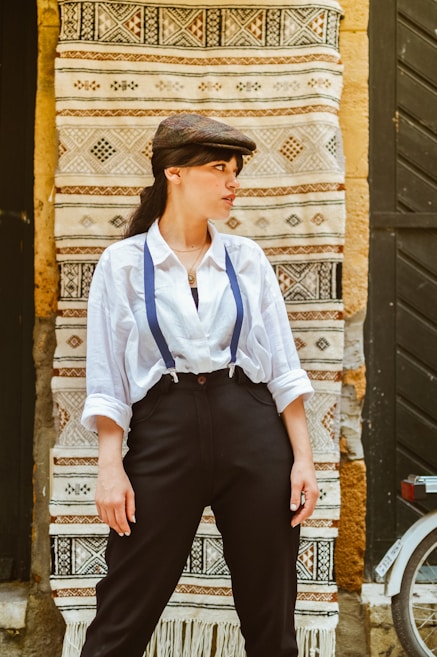 A person wearing a flat cap, white shirt, and black trousers stands in front of a patterned textile with geometric designs. The person appears to have one hand resting on their hip, and the textile includes colors like beige, brown, and black, creating an intricate backdrop.