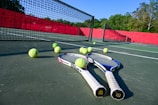 Tennis rackets and balls are scattered on a hard court with a net in clear view. The court is surrounded by a red barrier and lush green trees in the background, under a bright blue sky, creating a vibrant sports environment.