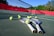 Tennis rackets and balls are scattered on a hard court with a net in clear view. The court is surrounded by a red barrier and lush green trees in the background, under a bright blue sky, creating a vibrant sports environment.
