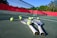 Tennis rackets and balls are scattered on a hard court with a net in clear view. The court is surrounded by a red barrier and lush green trees in the background, under a bright blue sky, creating a vibrant sports environment.