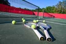 Tennis rackets and balls are scattered on a hard court with a net in clear view. The court is surrounded by a red barrier and lush green trees in the background, under a bright blue sky, creating a vibrant sports environment.