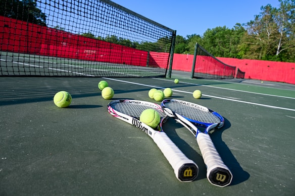 Tennis rackets and balls are scattered on a hard court with a net in clear view. The court is surrounded by a red barrier and lush green trees in the background, under a bright blue sky, creating a vibrant sports environment.