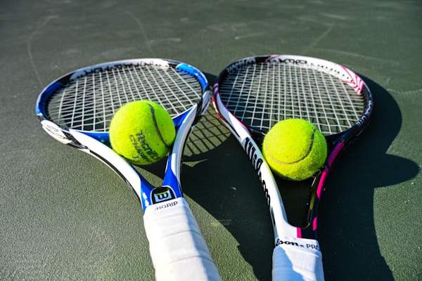 Two tennis rackets are lying on a court with tennis balls placed near the center of each racket. The rackets have blue and pink details with white grips, and the tennis balls are bright yellow.