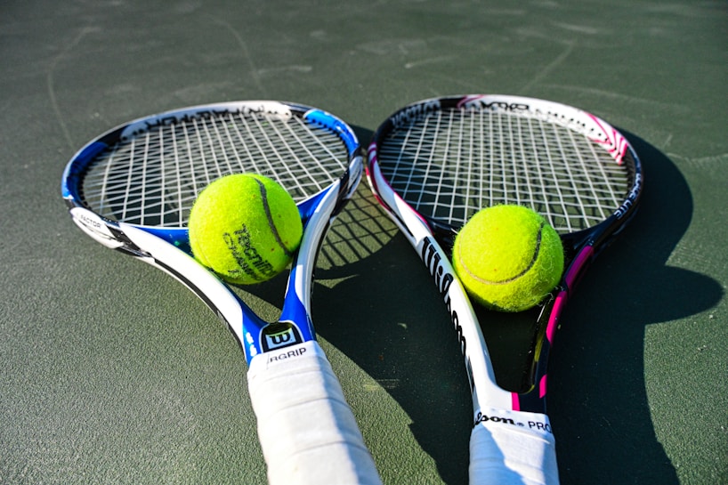 Two tennis rackets are lying on a court with tennis balls placed near the center of each racket. The rackets have blue and pink details with white grips, and the tennis balls are bright yellow.
